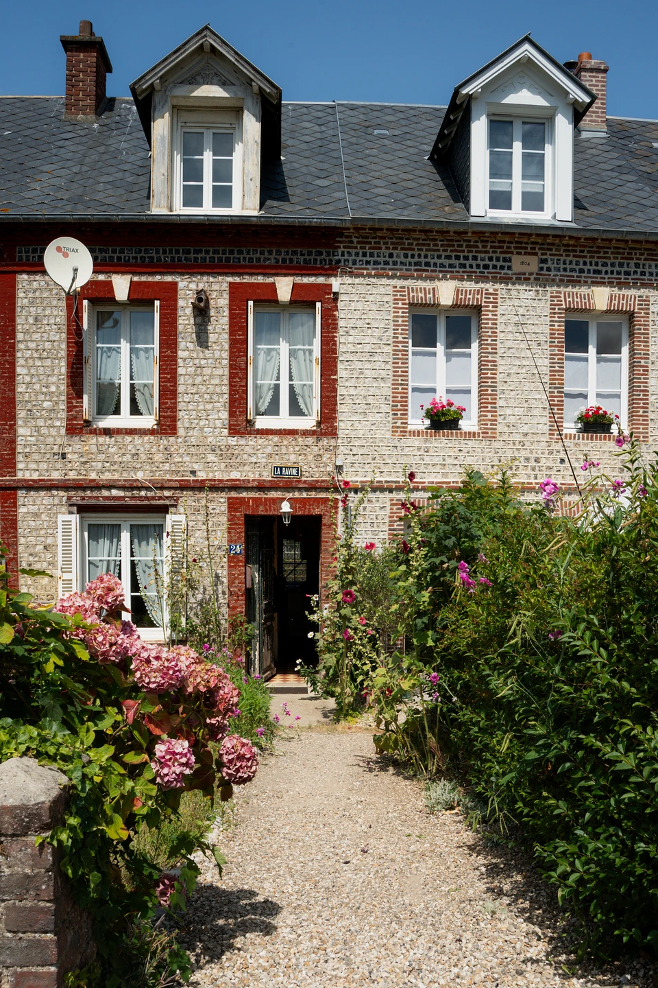 A stone house trimmed in red brick, a gravel path leading past hydrangeas and flowering shrubs