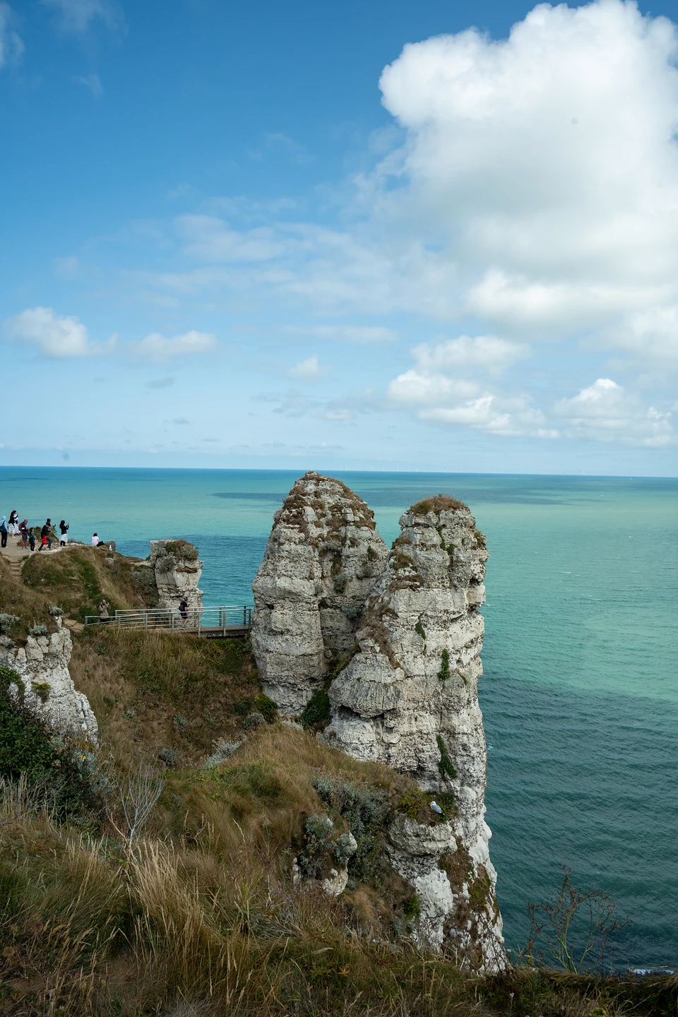 Two chalk pinnacles beside the cliff path, people at a railed viewpoint above turquoise sea