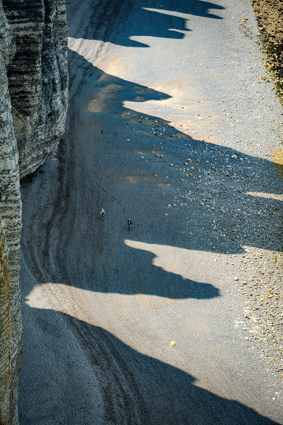 Jagged shadows of the cliff falling across a pebble beach, two walkers far below