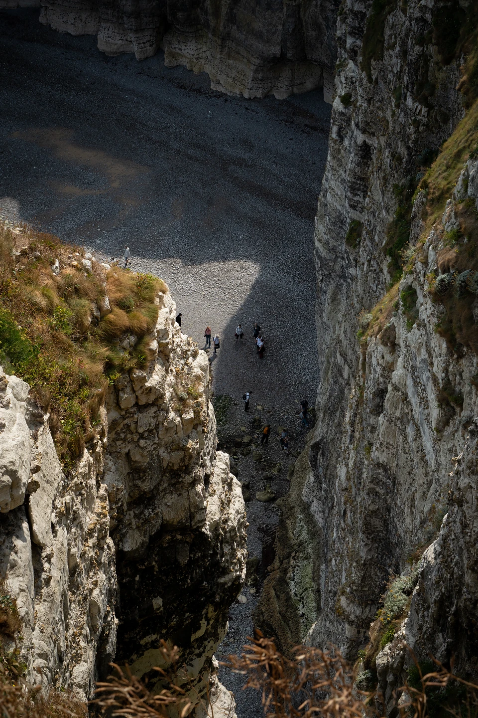 A narrow chalk gully dropping to a shaded pebble beach with small groups of walkers