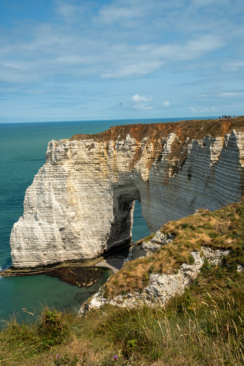 A massive layered chalk arch with a sliver of beach beneath, walkers on the clifftop beyond