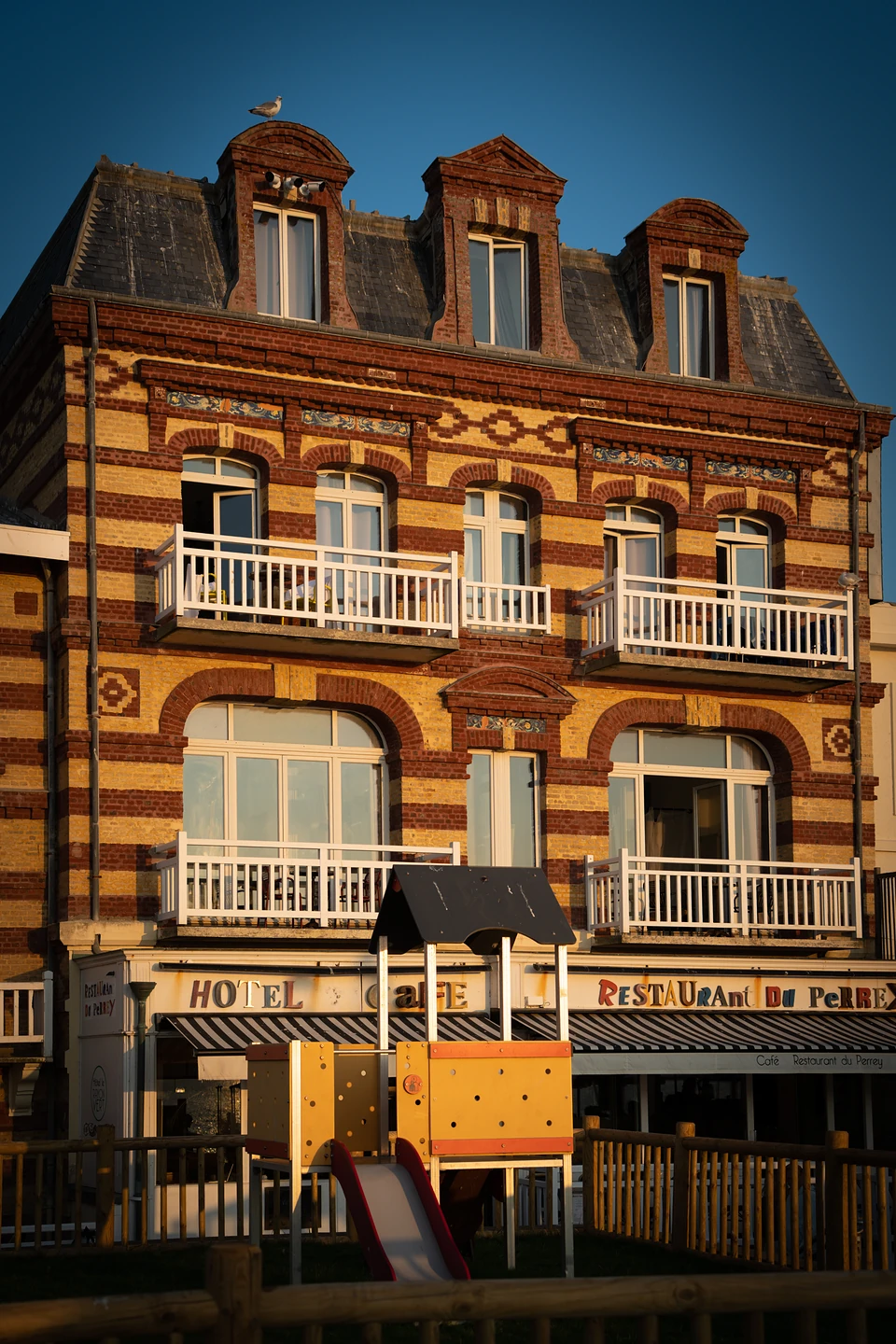 A striped brick hotel with white balconies in golden light, a children's slide in front