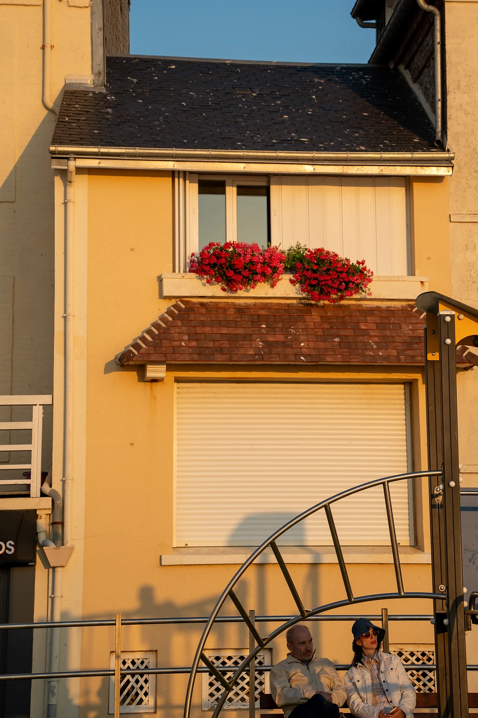 Red flowers in a window box on an ochre house, two people on a bench below