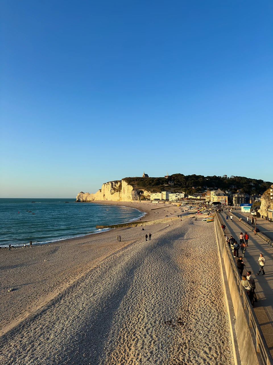 Low evening sun on the pebble beach and promenade, people strolling toward the far cliff