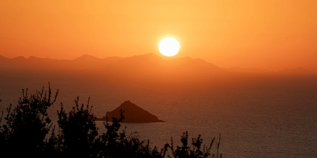 The sun sinking towards hazy mountains over the sea, a small pointed island in silhouette