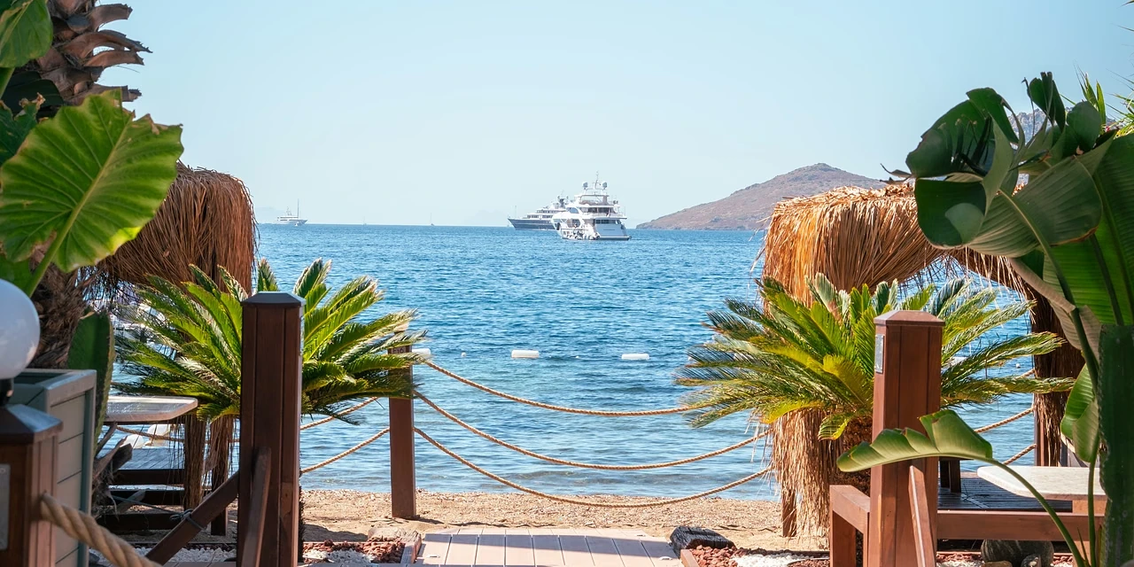 A boardwalk between rope rails leading to the beach in Yalıkavak, a yacht anchored offshore