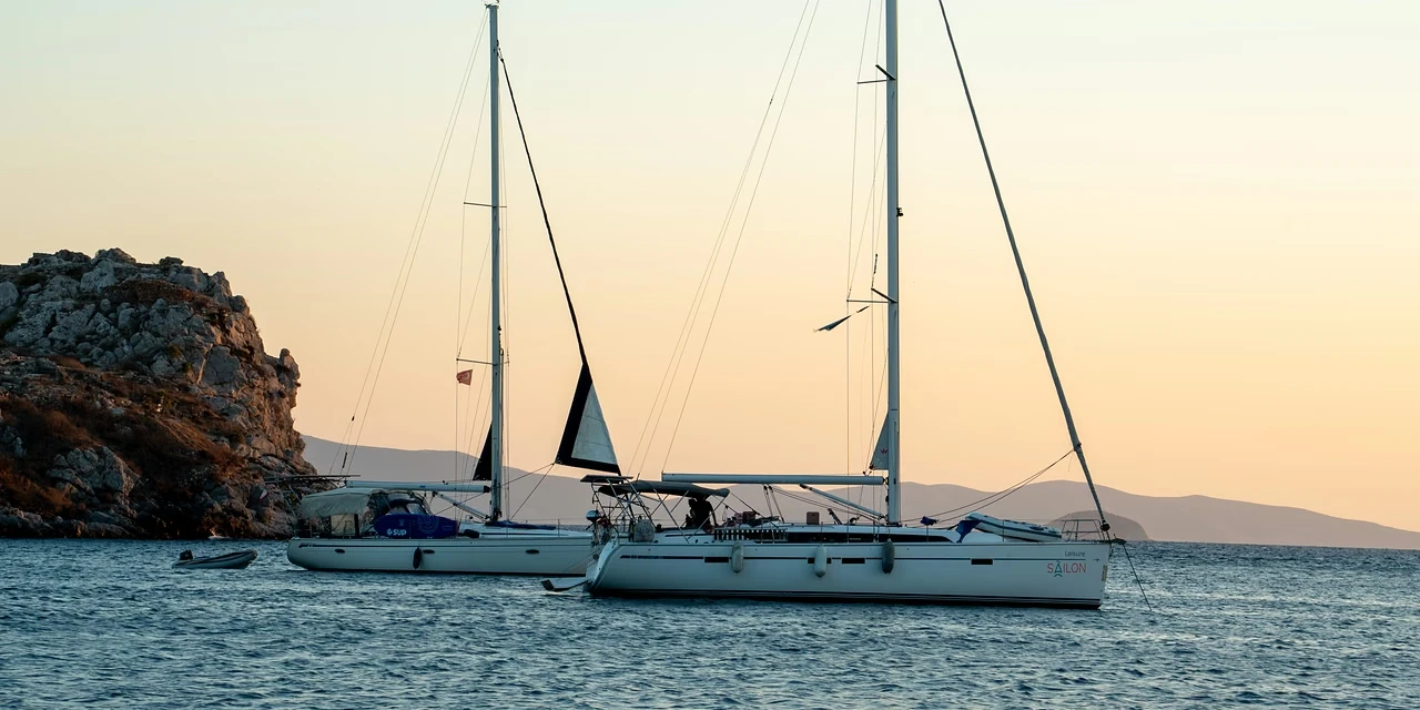 Two sailing yachts at anchor off a rocky headland in Gümüşlük, the dusk sky turning peach