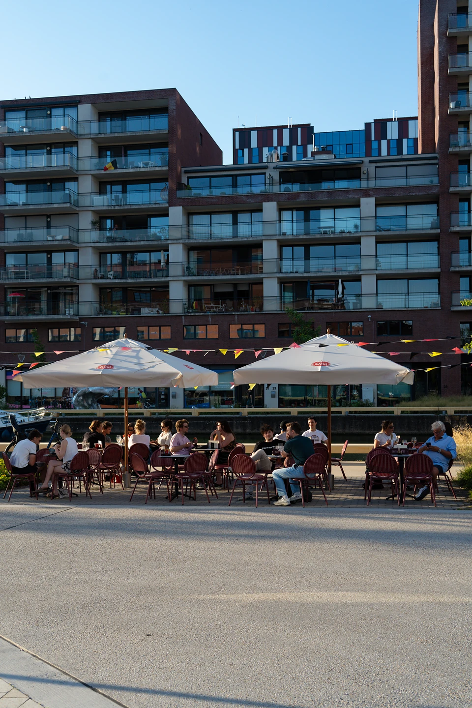People on red chairs under white parasols at a waterside terrace, apartment blocks behind