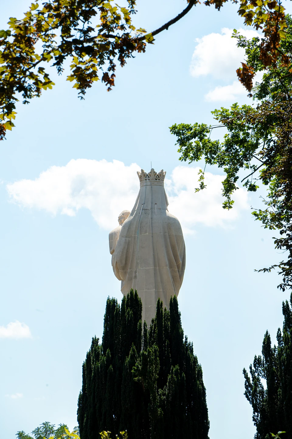 A white statue of a crowned figure holding a child, seen from behind above dark conifers