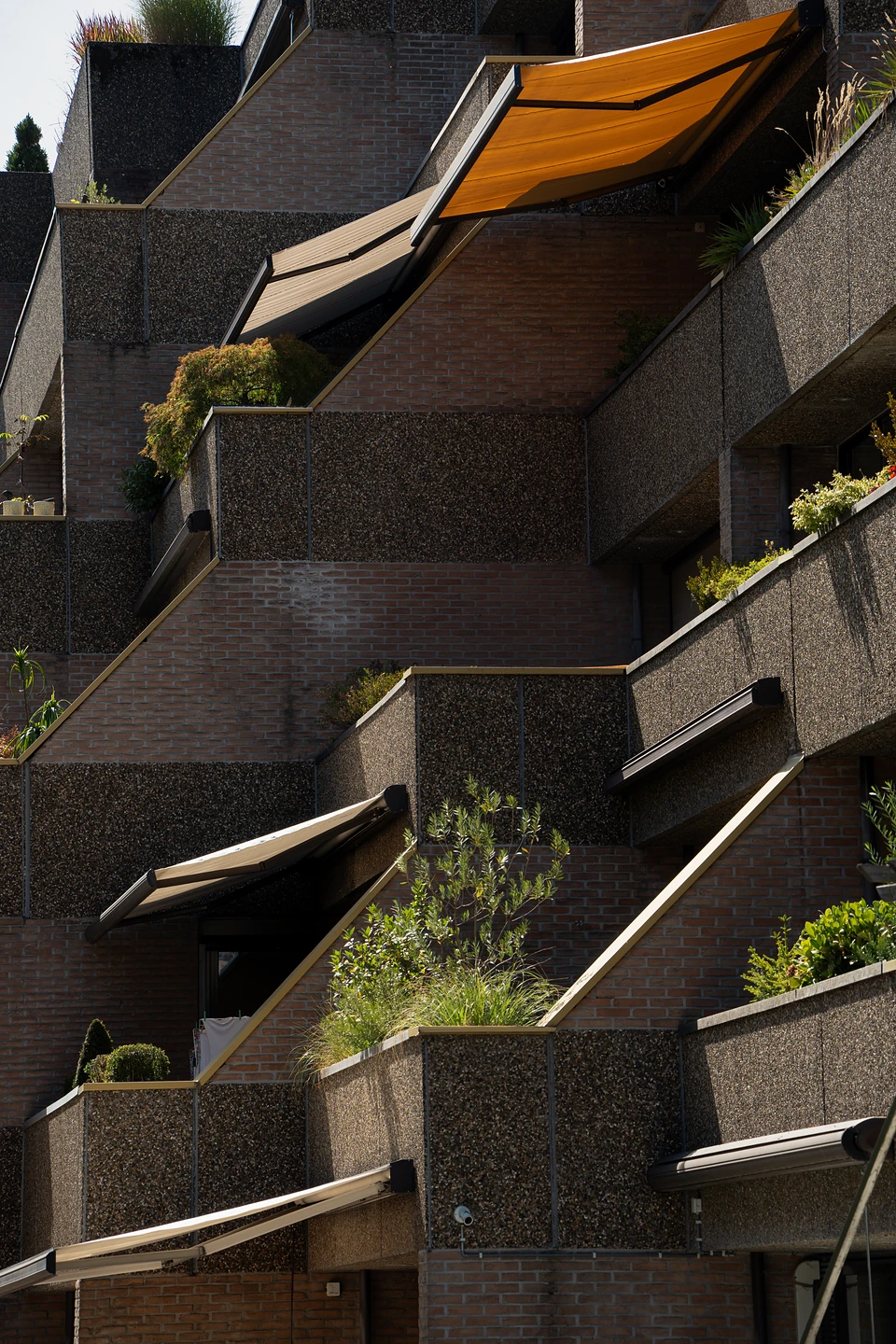 Stepped balconies of brick and pebbled concrete, with planters and an orange awning in strong sun