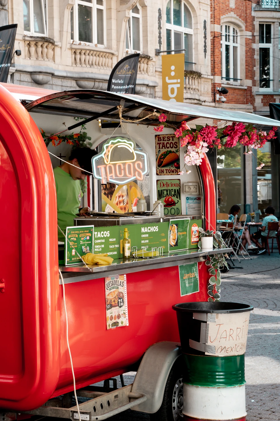 A red taco trailer trimmed with pink flowers, a ‘Tacos’ neon sign glowing in its hatch