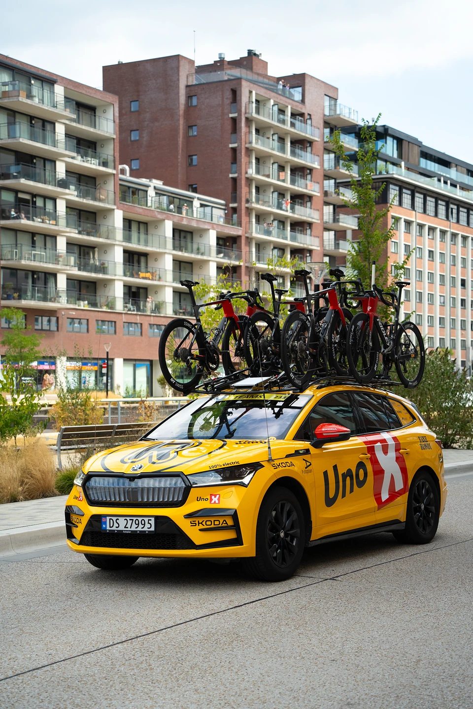 A yellow Uno-X team car with racing bikes on its roof, parked below apartment blocks