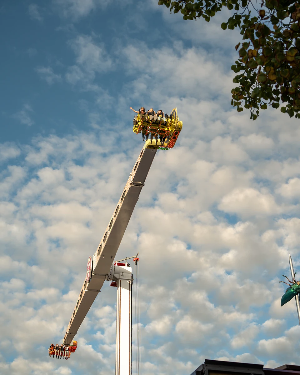 Riders at the top of a long swinging arm ride, held high against a cloud-flecked sky