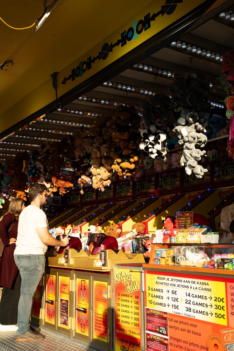 A man playing a ‘Flying Balls’ game while a woman watches, plush prizes hanging overhead