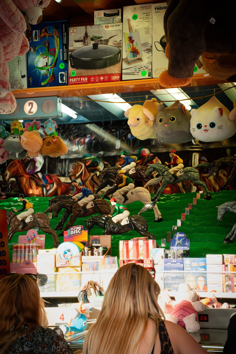 Toy jockeys on a green horse-race game, plush prizes above and two players at the counter