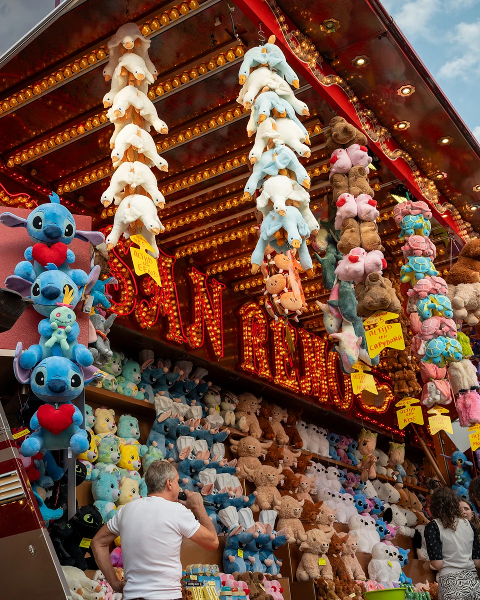 A man at a prize stall counter, towers of plush toys hanging from the bulb-lit roof