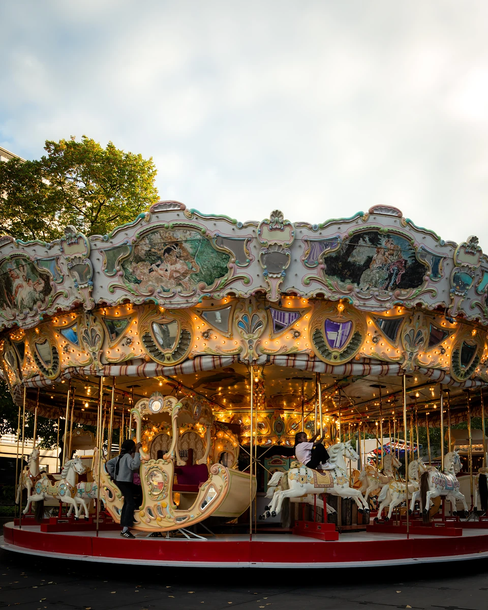 The whole carousel lit up beneath a pale evening sky, painted scenes around its crown