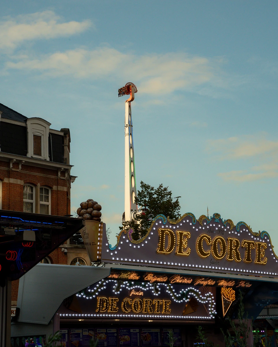 The bulb-lit sign of the De Corte snack stall, a ride’s long arm standing upright behind