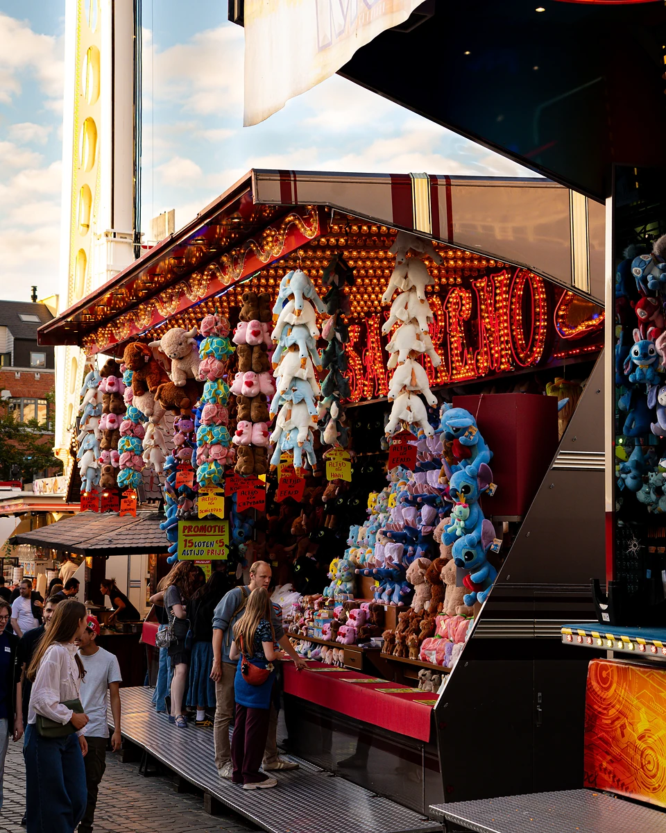 Visitors browsing a plush-toy stall in late sun, a ride’s white arm rising behind