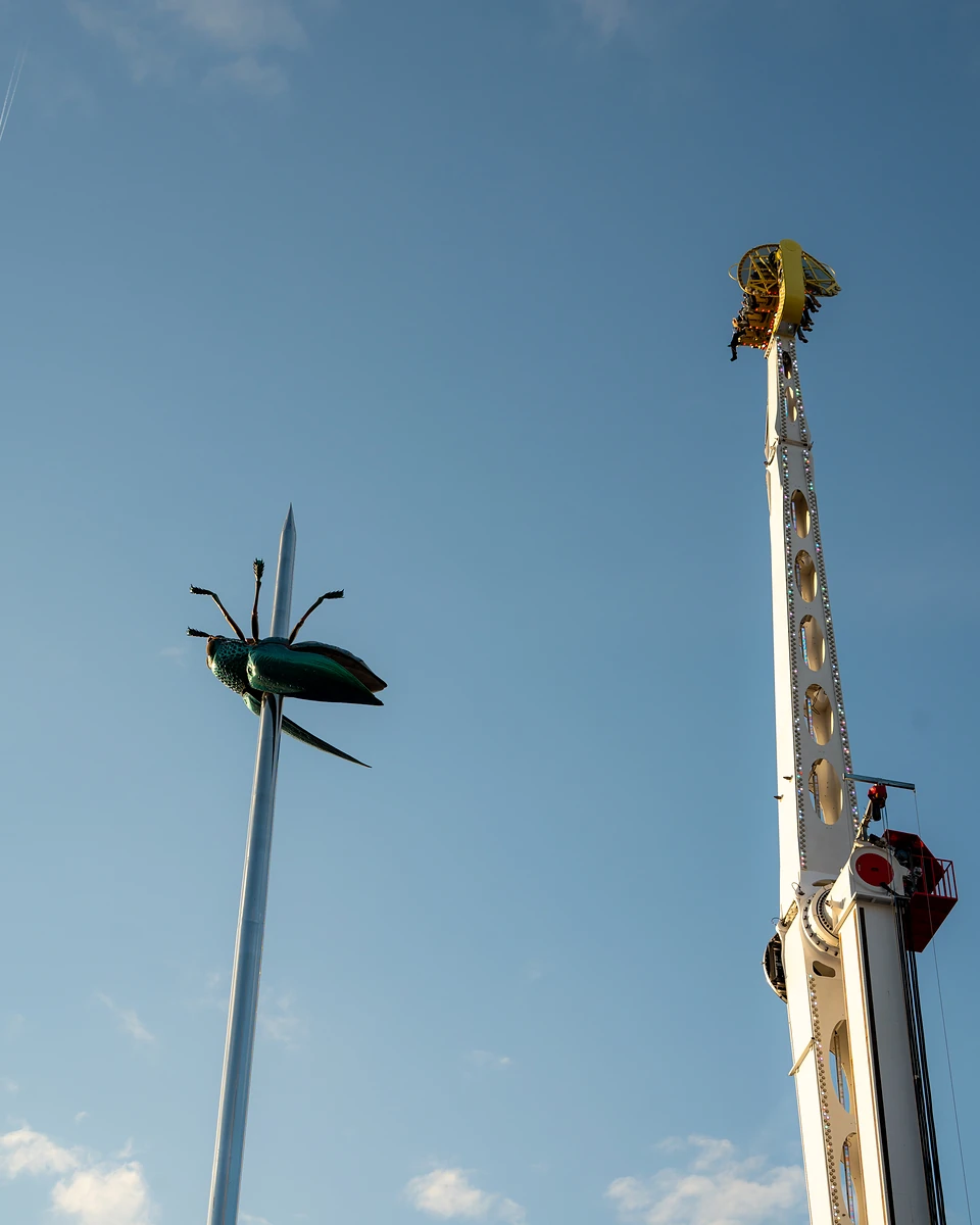 The giant green beetle on its needle beside a ride’s upright arm, riders at the top