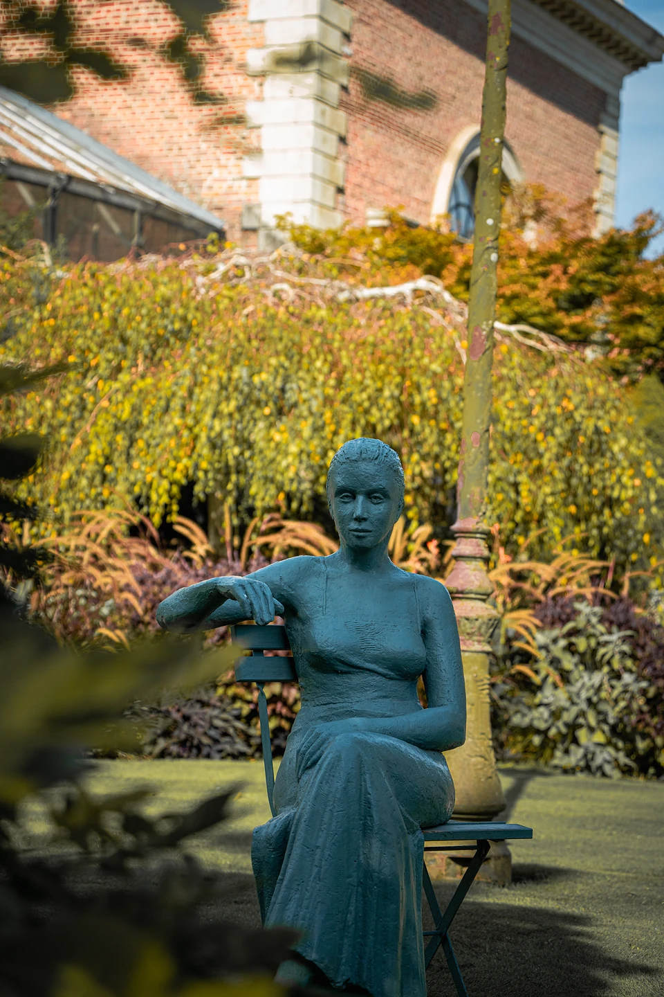 A blue-green sculpture of a woman seated on a garden chair, a red-brick building behind