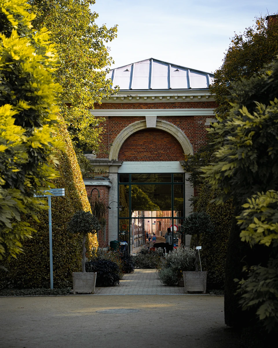 A glass-roofed brick entrance at the end of a hedged path, the street glimpsed through it