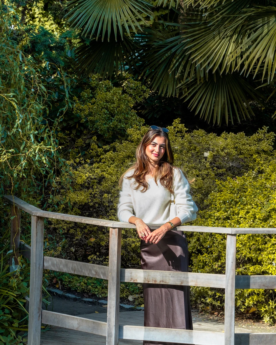 Ipek in a cream jumper leaning on a wooden footbridge railing, palm fronds above