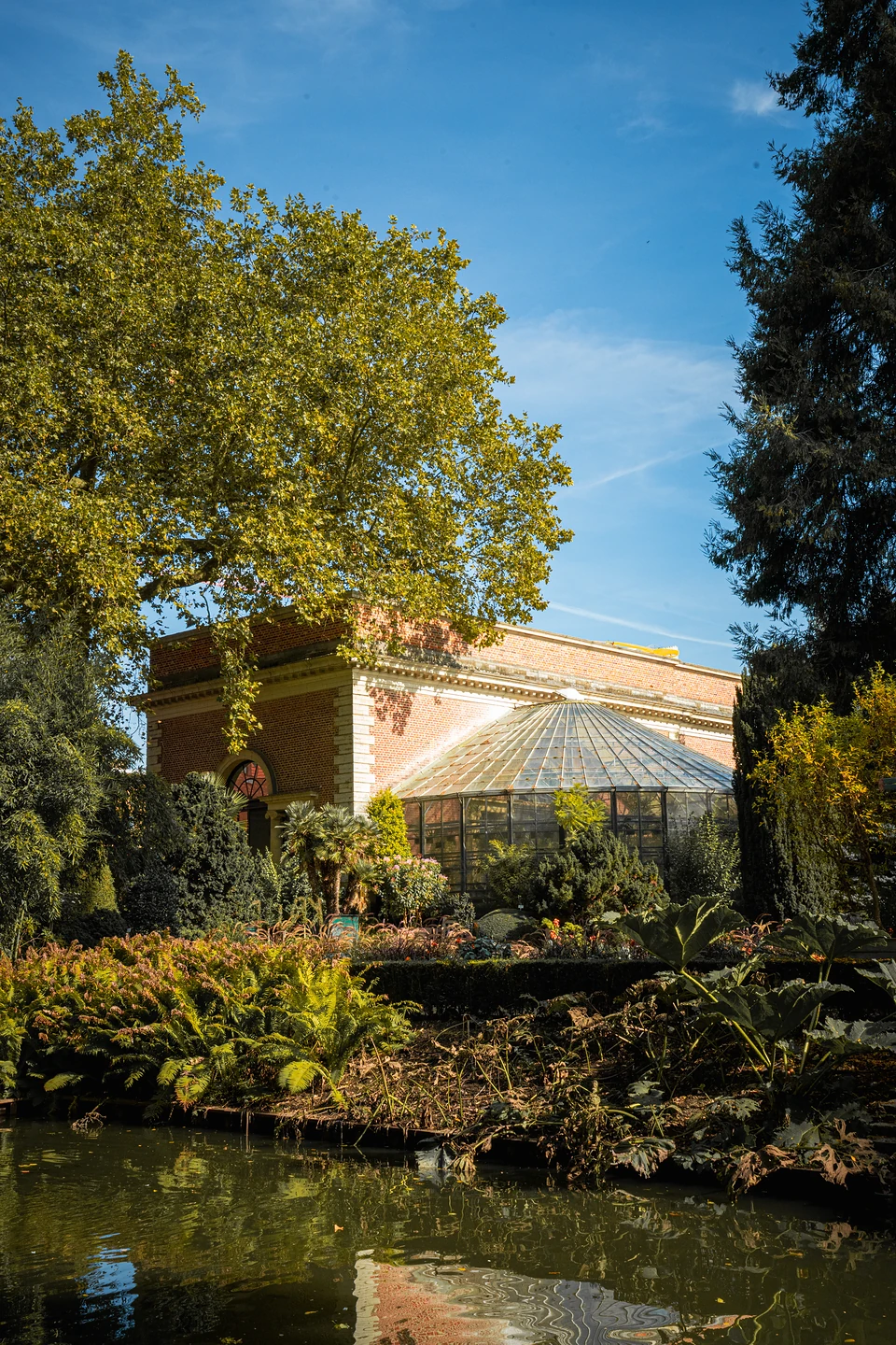 A domed glasshouse and red-brick pavilion seen across a pond under a blue sky