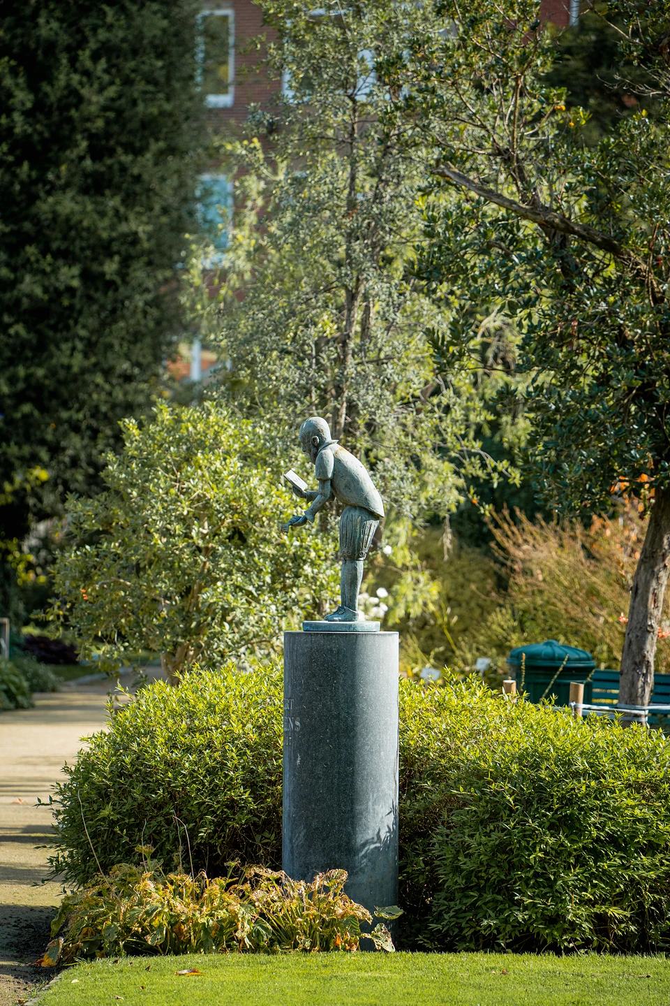 A small statue of a child reading, on a tall stone column among clipped shrubs