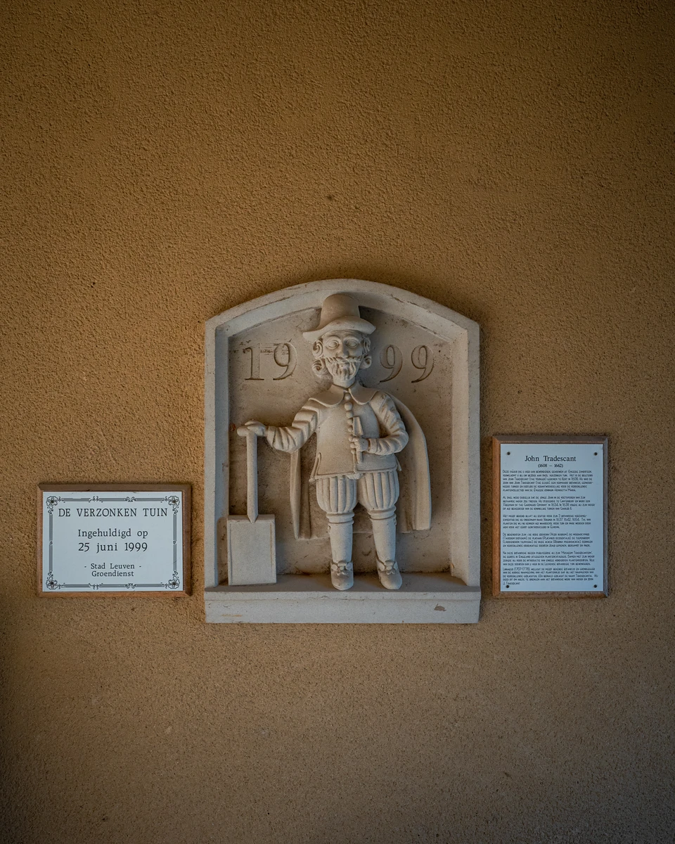 A stone relief of a figure holding a spade, beside a plaque reading “De Verzonken Tuin”