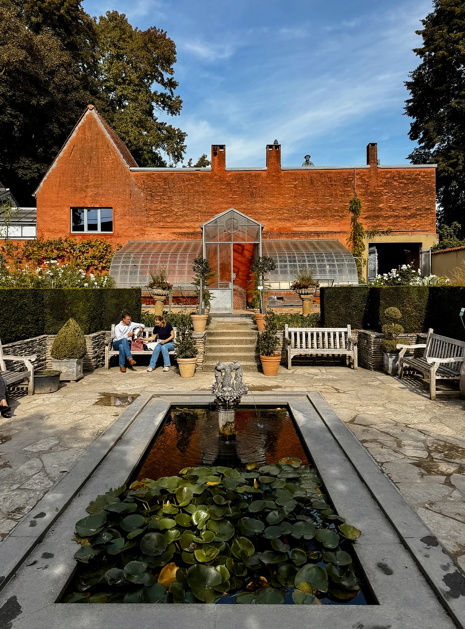A lily pond and fountain before a brick building and glasshouse, two people on a bench