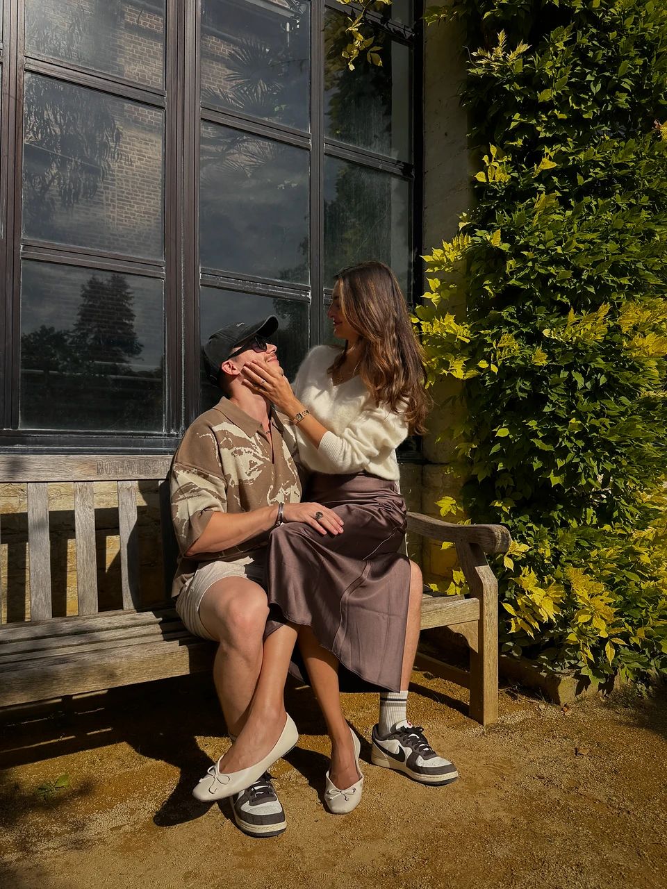 Ipek and Robbe close together on a wooden bench beside tall glasshouse windows, in low sun