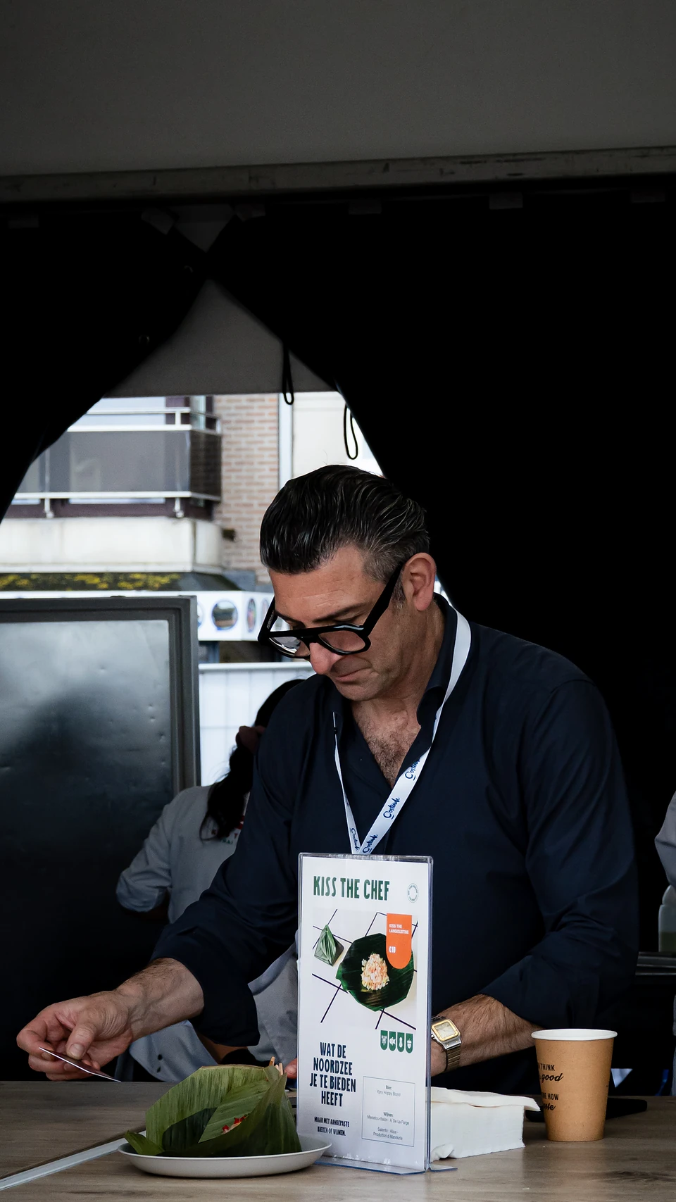 A man in black-rimmed glasses at a 'Kiss the chef' stand, a leaf-wrapped dish in front