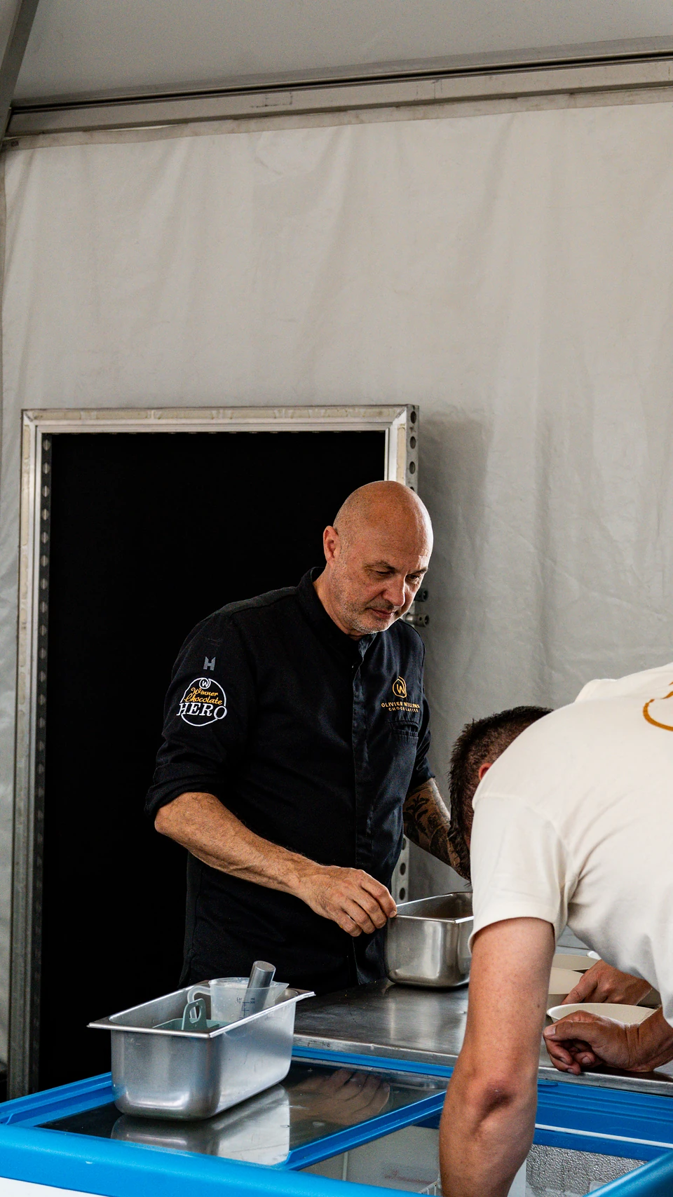 A chef in a black jacket working at a steel counter in a festival tent