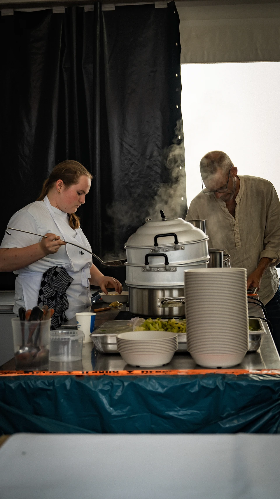 A cook in whites ladling beside a tall stacked steamer, steam rising past a second cook