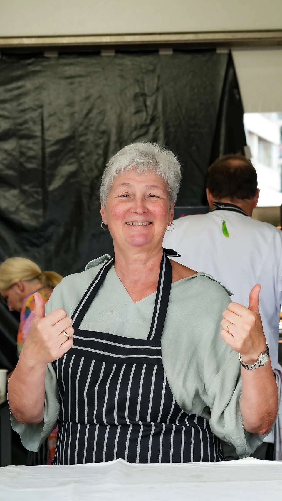 A woman in a striped apron smiling and giving two thumbs up at her festival stand