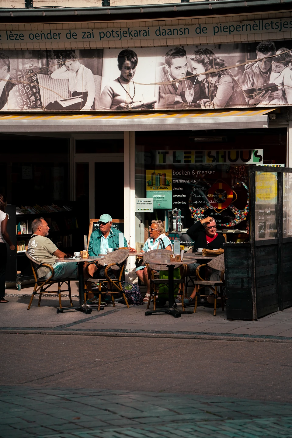 People at sunlit terrace tables outside the Leeshuus, old black-and-white photos printed above