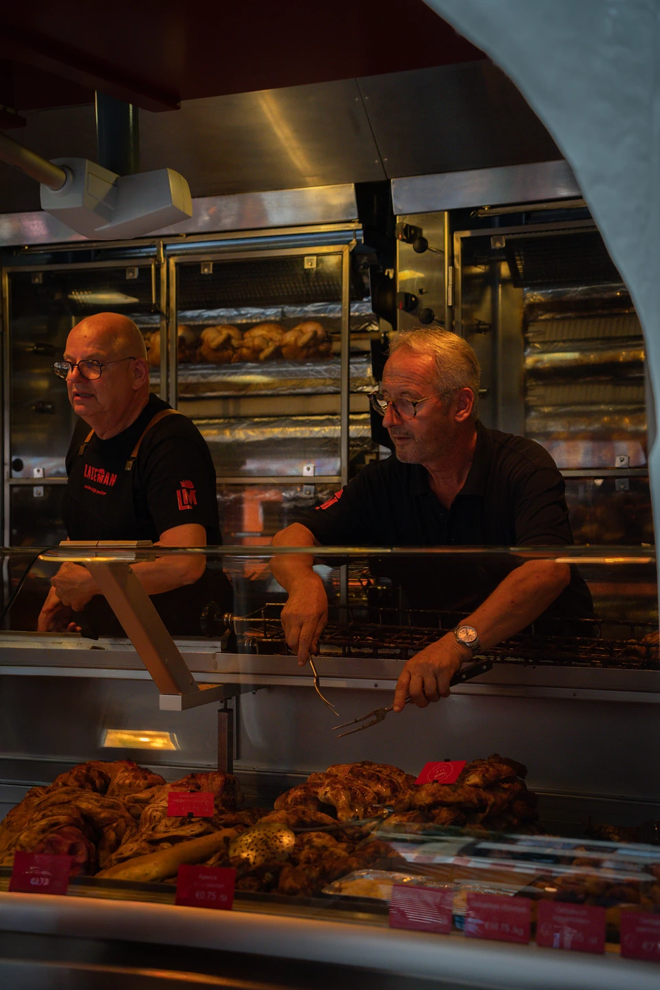 Two men in black at a rotisserie counter, roast chickens in the ovens at their backs