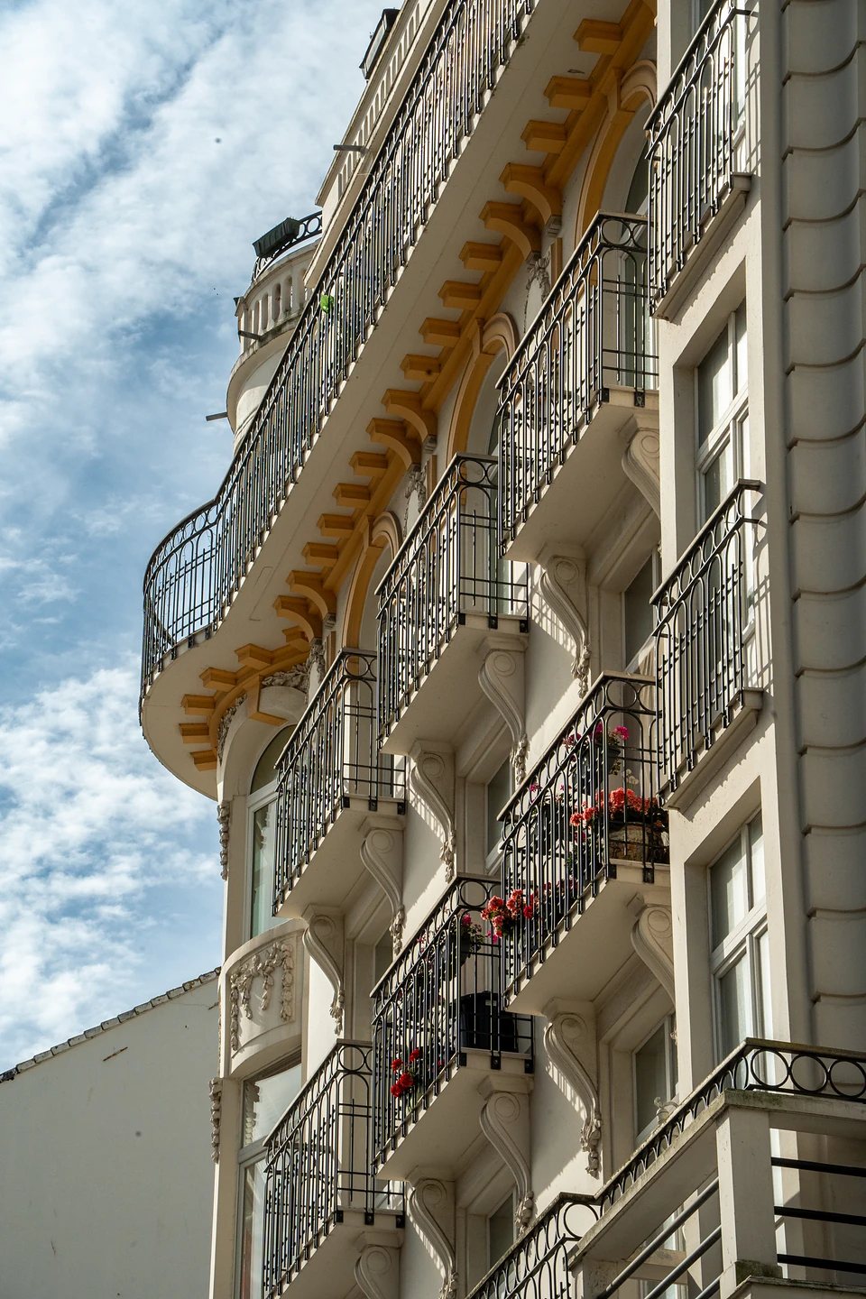 Curving wrought-iron balconies with red flowers on a white facade, ochre brackets under each tier