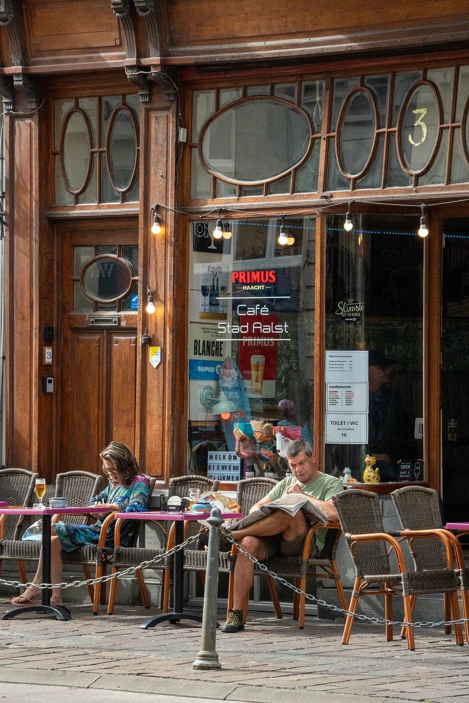 Two people on the wicker terrace of Café Stad Aalst, one reading a newspaper