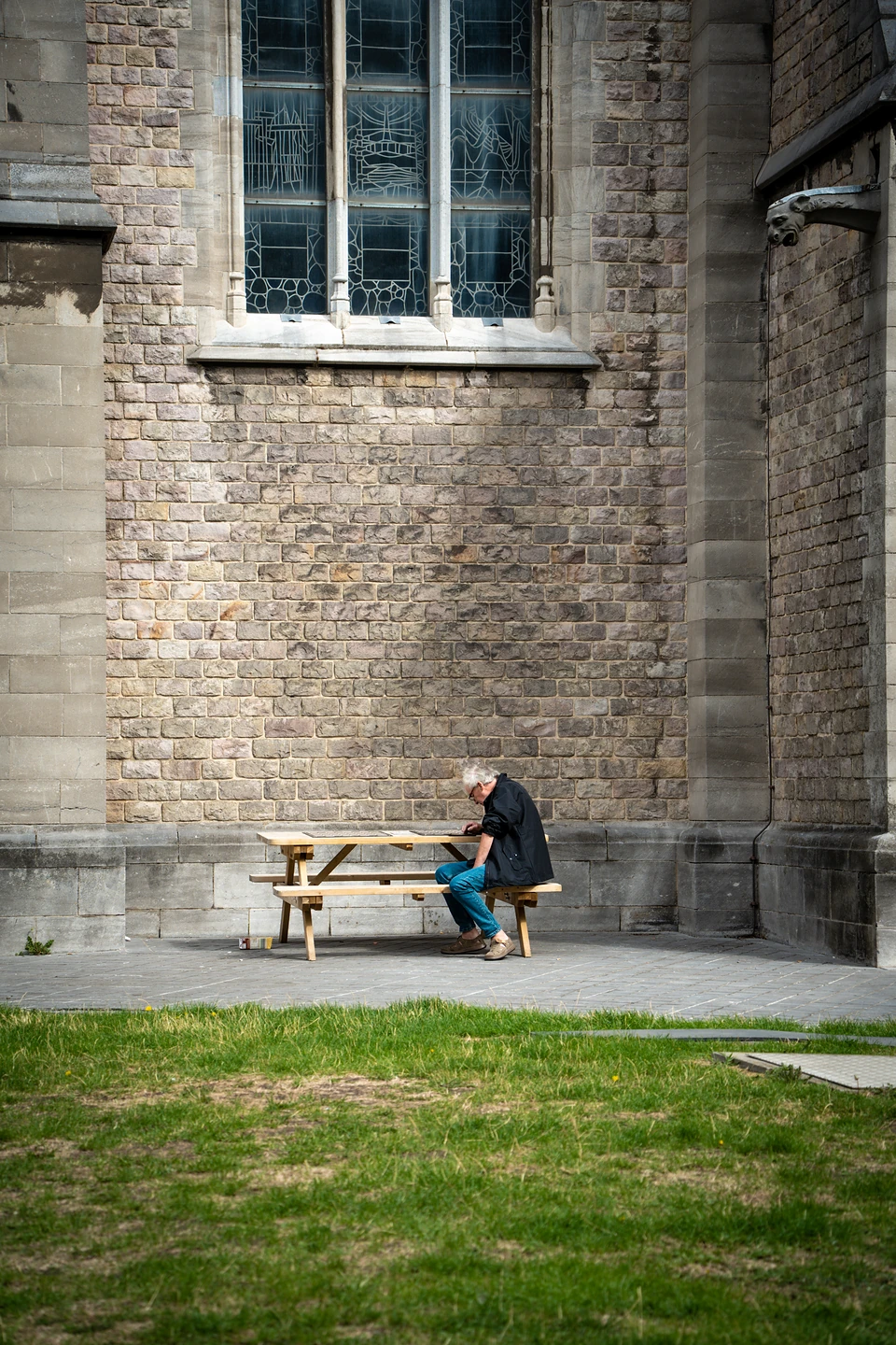A man alone at a picnic table against a stone church wall, below a stained-glass window