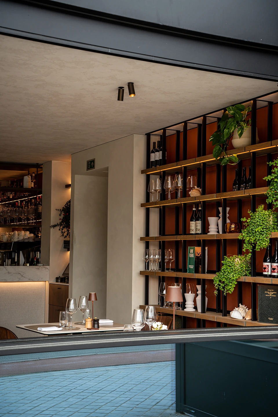 Lit shelves of wine bottles, glasses and trailing plants beside a laid table in a restaurant