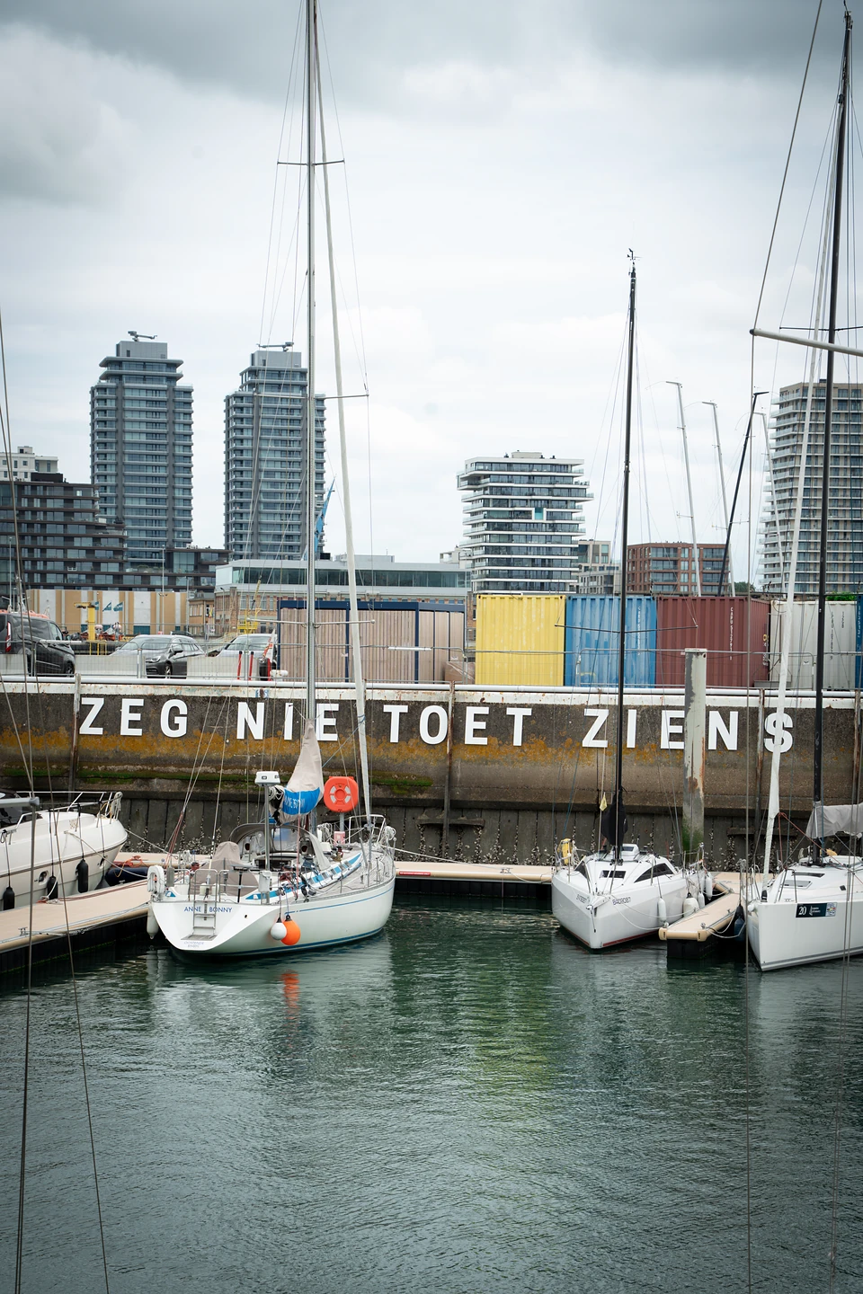 Sailboats moored below a quay wall lettered 'Zeg niet toet ziens', tower blocks behind