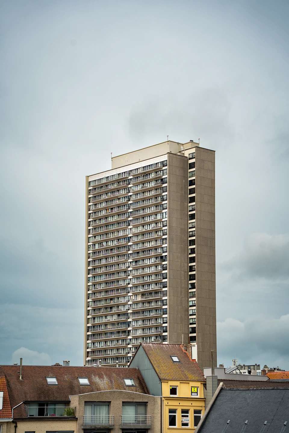 A tall apartment tower rising above tiled rooftops under a heavy grey sky