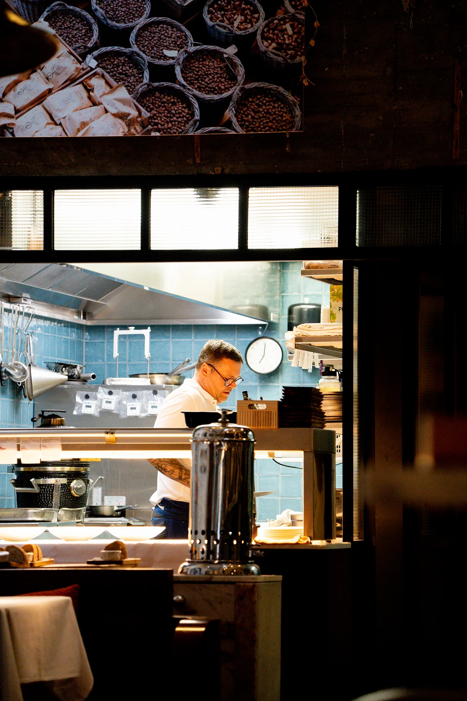 A cook in a white shirt at work behind the pass in the blue-tiled open kitchen
