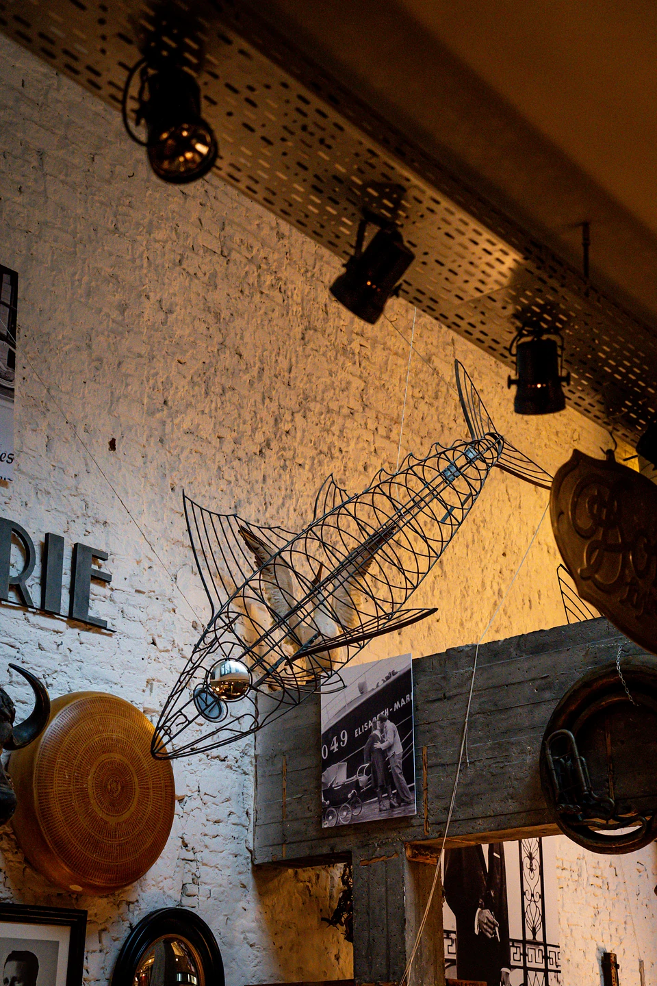 A large wire fish sculpture hanging under spotlights against the whitewashed brick wall