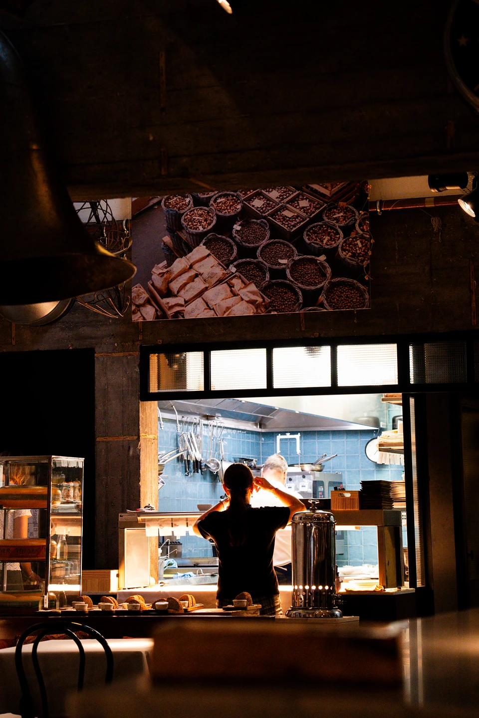 A server silhouetted at the lit pass, a cook at work in the blue-tiled kitchen beyond