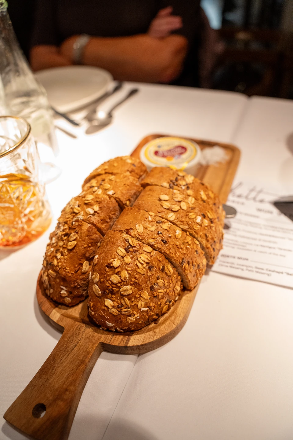 A sliced seeded loaf on a wooden board, with butter and salt, beside the menu