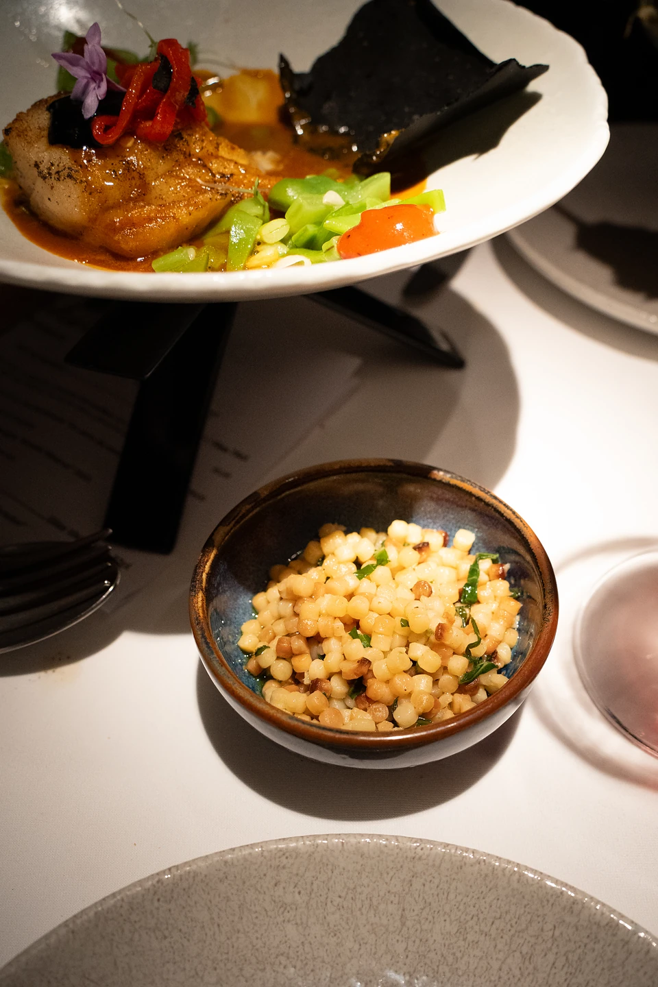 A side of small pasta pearls with herbs in a glazed bowl, the fish course behind