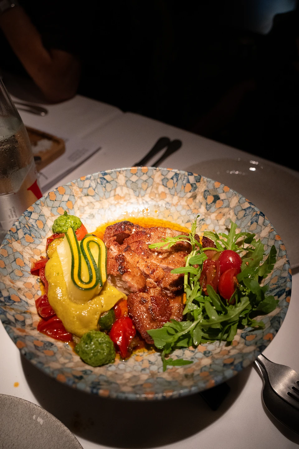 Sliced grilled meat with rocket, roast tomatoes and a yellow purée in a mosaic-patterned bowl